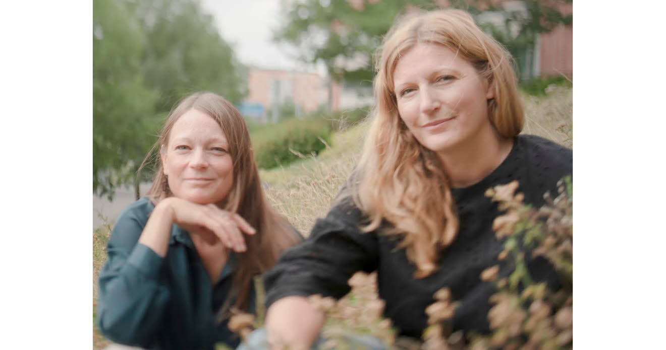 Foto på Jenny och Lotta som sitter ner i gräset, i bakgrunden skymtas träd och en tegelbyggnad.