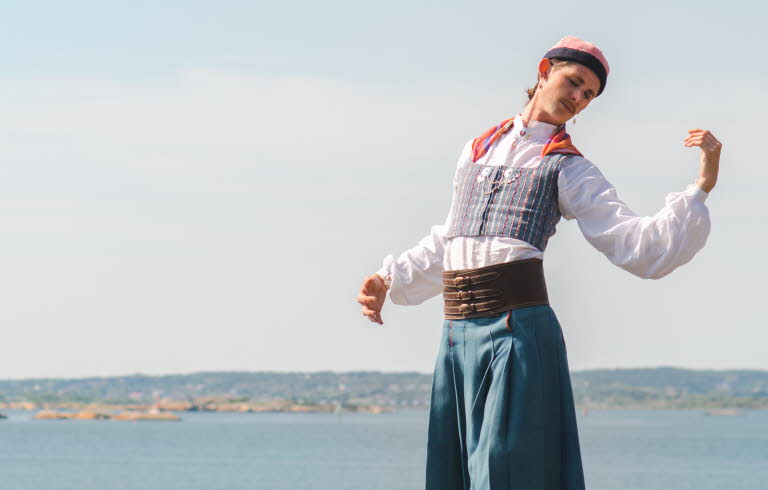 Person i folkdräkt står på en klippa vid havet med öar i bakgrunden.