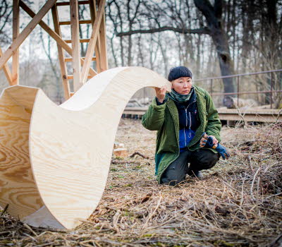 En kvinna som sitter utomhus lyssnar i en överdimensionerad tratt gjord av trä.