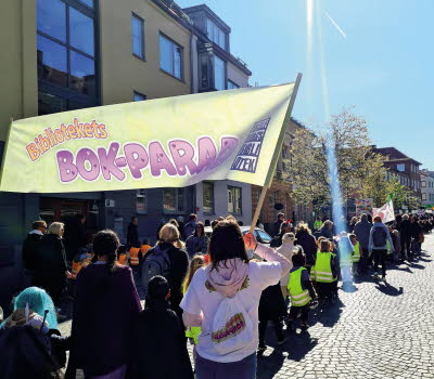 Barn och vuxna som tågar längs en gata med banderoller med texten Bibliotekets bokparad.