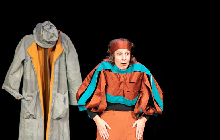 Två kvinnor på scen. En spelar på keyboard som ligger på ett figurliknande träbord och den andra tittar storögt mot publiken, axlarna är upphöjda. Båda har färgstarka kläder i turkost och ockra. I bakgrunden hänger en grå rock med tillhörande hatt.