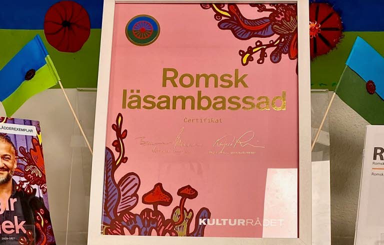 Hylla från bibliotek med intyg på att biblioteket är romsk läsambassad.