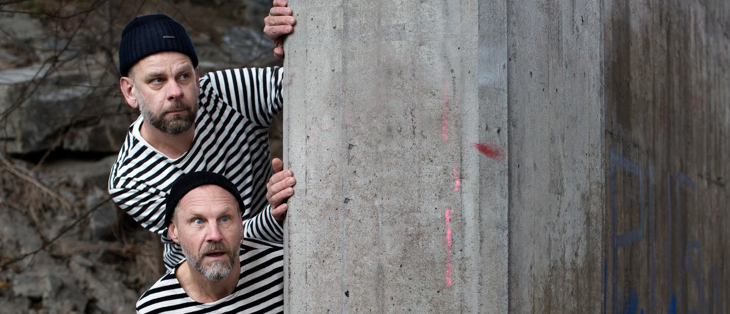 Anders Sparring and Per Gustavsson, dressed in black and white sweaters/prison style, looking out behind a wall.