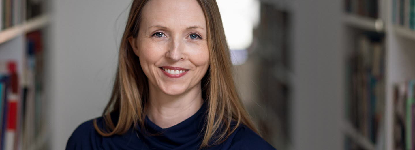 Medelålders kvinna med långt cendréfärgat hår och blå tröja omgiven av bokhyllor.