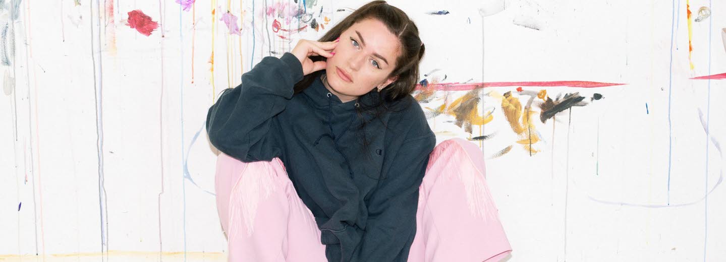 Artist Moa Romanova is sitting against a wall in pink pants and a black hoodie.