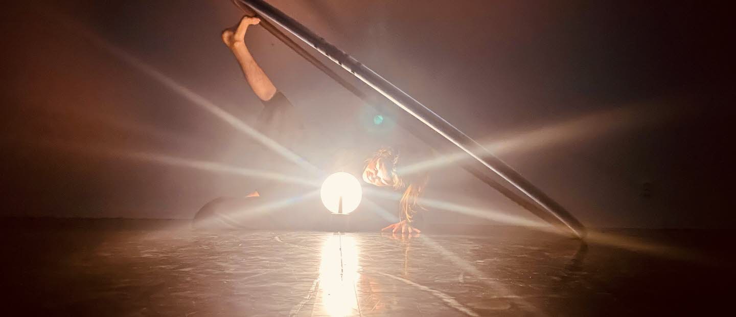 En kvinnlig dansare ligger på sidan i ett mörkt rum. Det ena benet är uppsträckt där foten balanserar en stor ring. Framför henne står en lampa som likt en strålkastare riktas mot betraktaren.