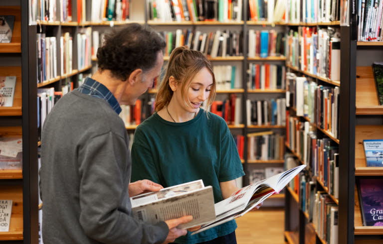 Två bibliotekarier står bland bokhyllor, bläddrar i varsin bok och diskuterar innehållet. 