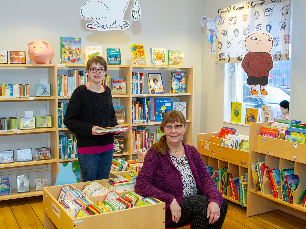 Två kvinnor, en sittande och en stående, står i bibliotekets barnboksområde. En bild av Alfons Åberg hänger vid fönstret.