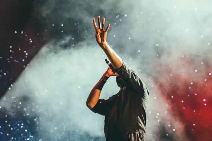 Man som sjunger i mikrofon med vänstra armen upp i luften. I bakgrunden syns rök i vitt och rött samt ljus som påminner om stjärnor.