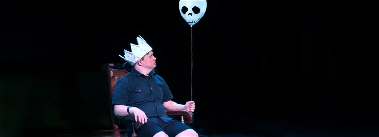 En man sitter på en scen. På huvudet har han en kungakrona i papp, i handen håller han en ballong med spökansikte.