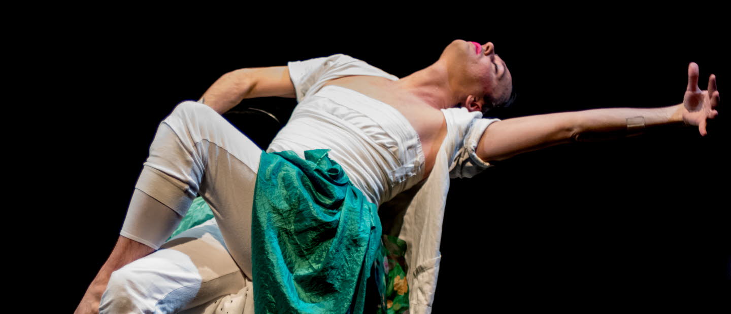 En dansare som sitter på en stol och gör en rörelse bakåt. Långa textiler som släpar på scengolvet. 