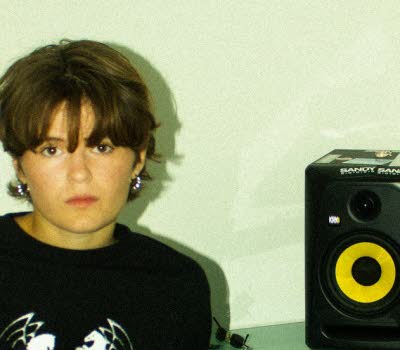 Kvinna i 20-årsåldern sittandes vid ett bord framför en laptop. På bordet en högtalare och hörlurar. 