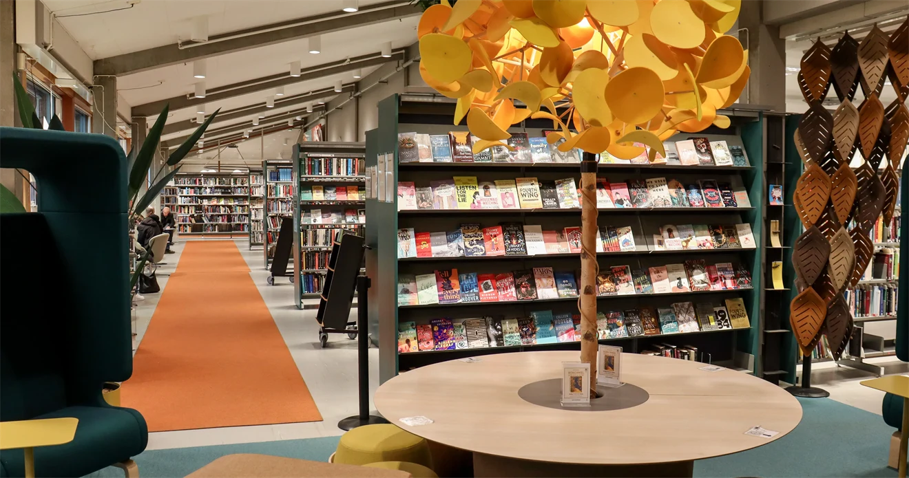 Interiör från bibliotek med bokhyllor, sittmöbler och samlingsbord med en lampa som ser ut som ett träd.