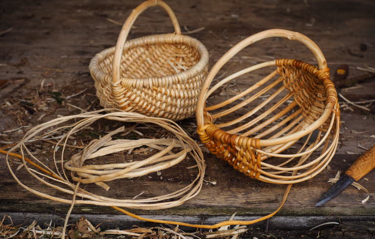 Två flätade korgar i naturmaterial, omgivna av rötter och en kniv på ett träbord.