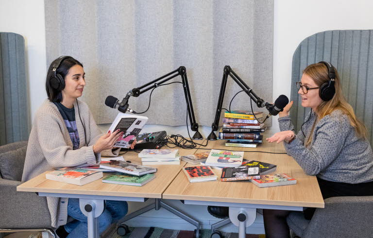 Två biblioteksmedarbetare sitter i en poddstudio med massa böcker som de diskuterar. 
