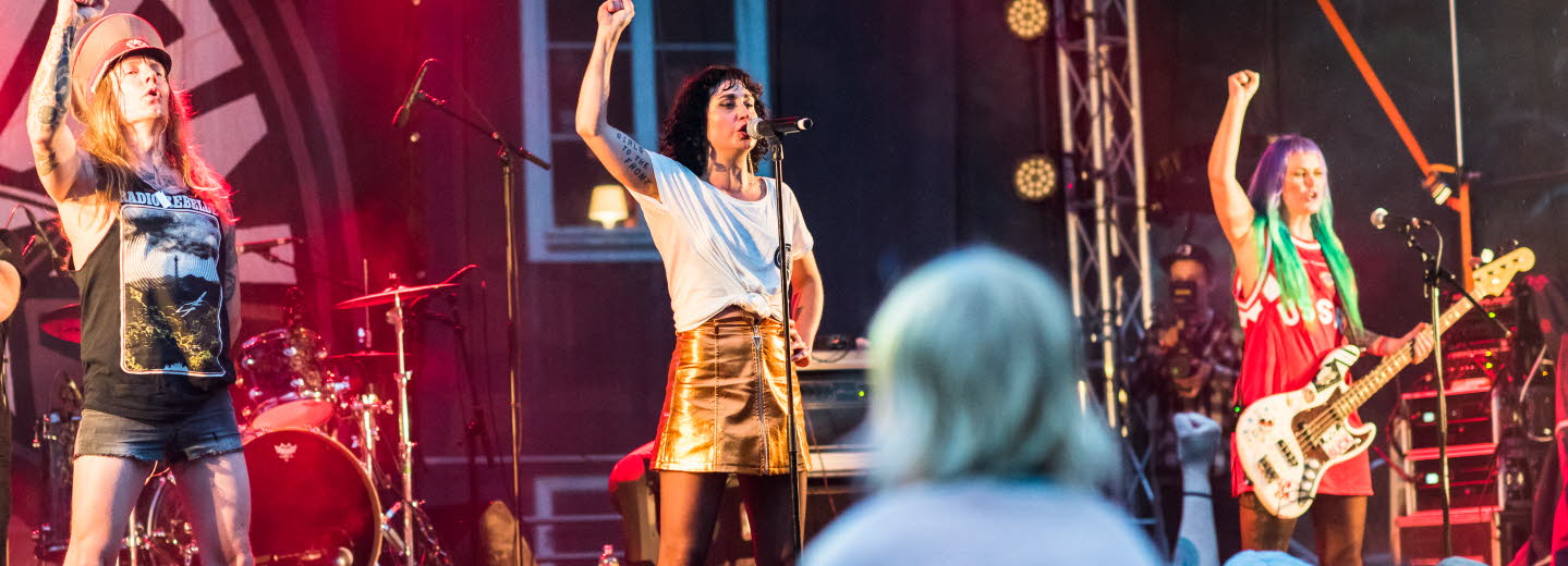 Three performers on a music stage raising their right hand and singing.