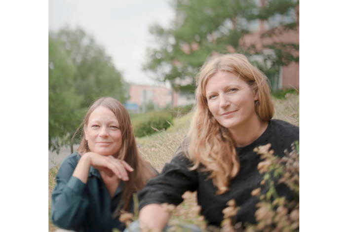 Foto på Jenny och Lotta som sitter ner i gräset, i bakgrunden skymtas träd och en tegelbyggnad.