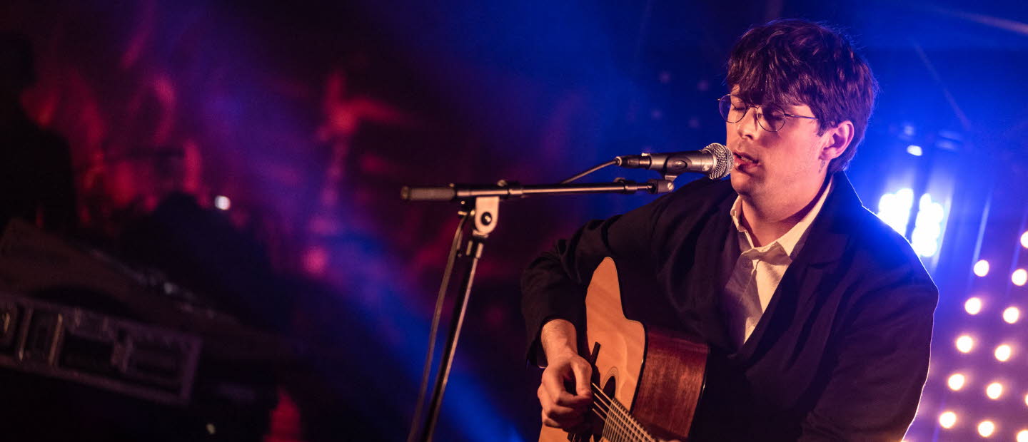 Ung man sitter på en scen, spelar gitarr och sjunger. 