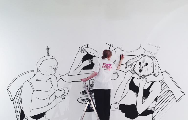 Bim Eriksson is painting on a wall. She's painting four women sitting att a dinner table.