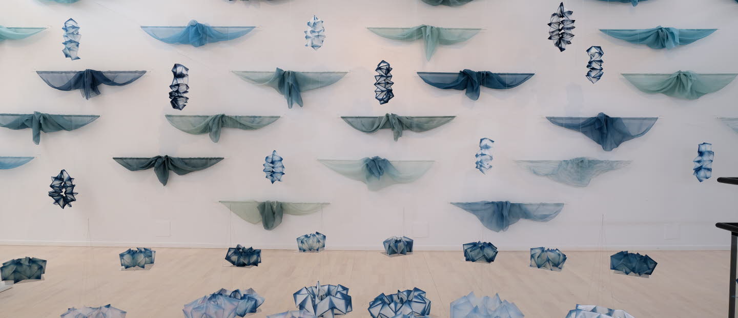 Konstinstallation med blå och gröna tygstycken upphängda i rader på en vit vägg, samt geometriska former i liknande färger placerade på golvet.