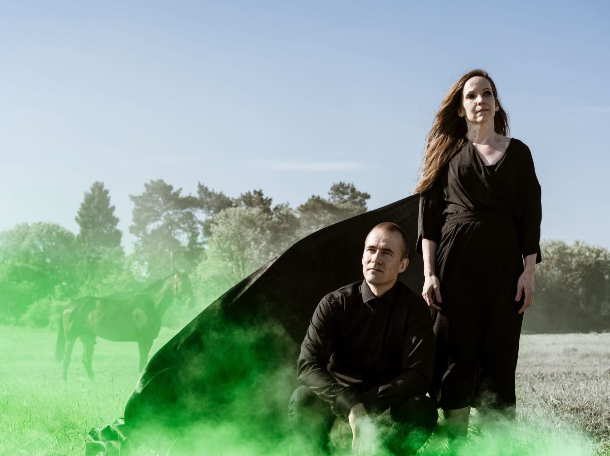 En man och en kvinna i svart klädsel på ett torrt gräsfält med grön rök.