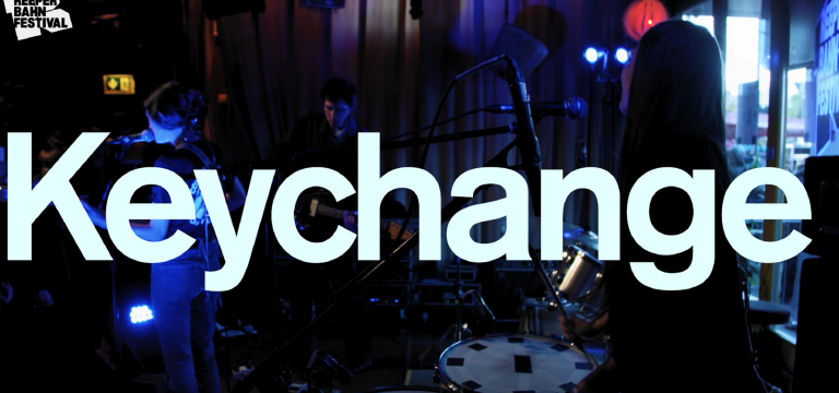 Vit text med Keychange, i bakgrunden ett band som spelar.