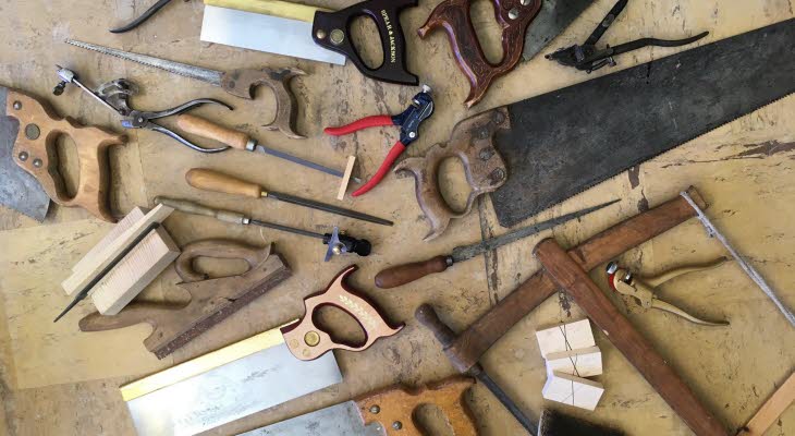 Gamla och nya handverktyg, främst sågar, filar och tänger, utspridda på ett slitet träbord.