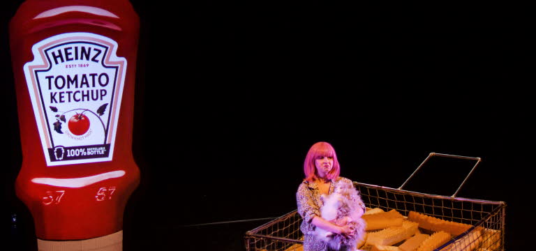En tjej i rosa hår står på en mörk scen, i bakgrunden syns en förstorad ketchupflaska och förstorade pommes frites..