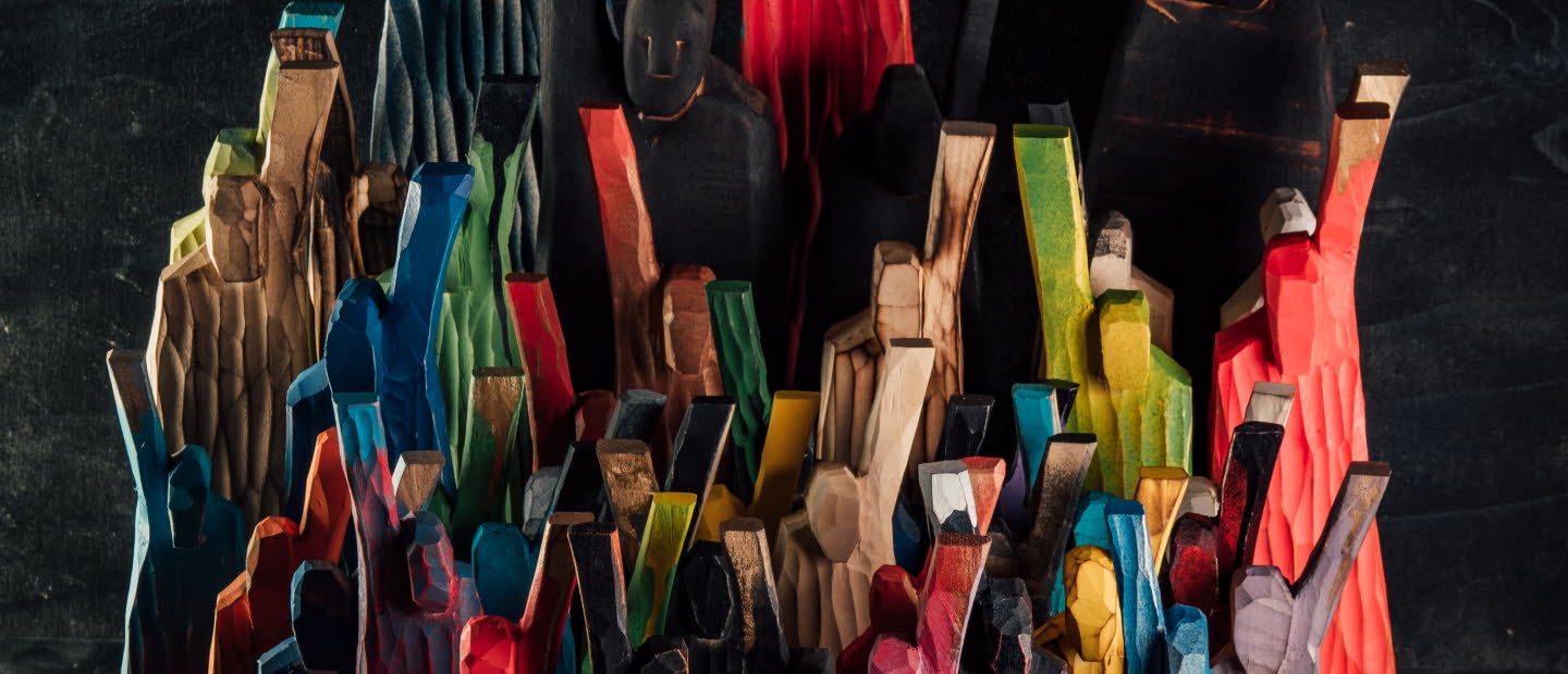 Flera färgglada, grovt täljda träfigurer står tätt tillsammans mot en mörk bakgrund, med armar lyfta i olika vinklar.