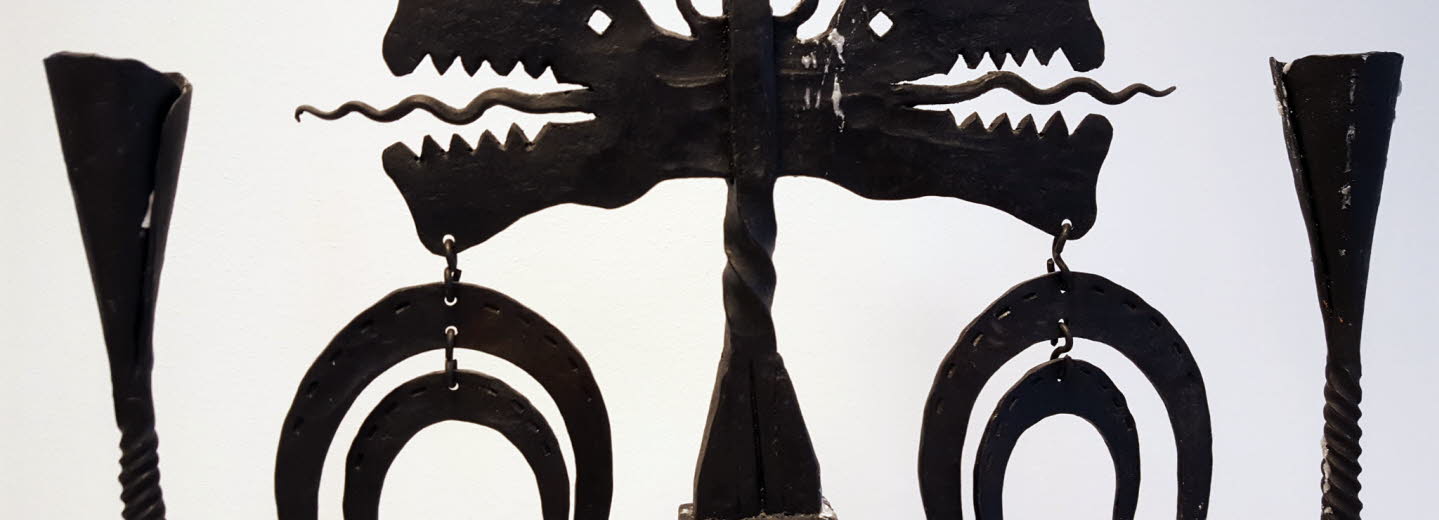 Överdelen av en ljusstake i smide med två drakhuvuden och hästskoformade hängen.