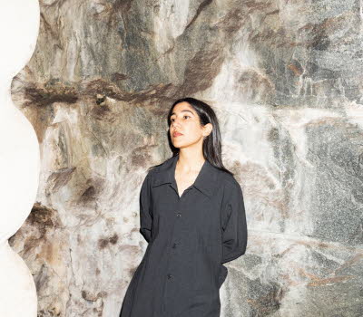 Leila is wearing black clothes and leaning to a stone wall.