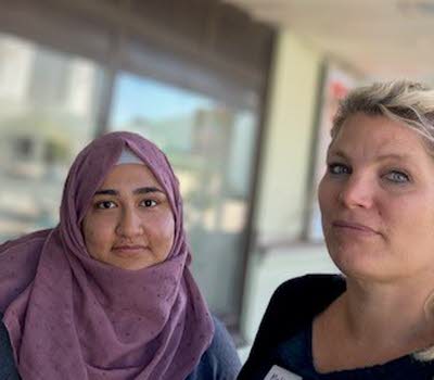 Två kvinnor står tillsammans utanför ett bibliotek, den ena bär en hijab, den andra har håret utsatt. 