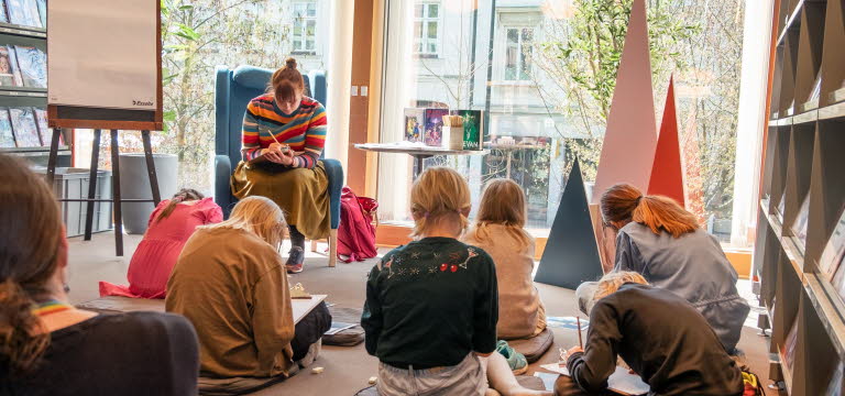 En grupp barn sitter utspridda framför en kvinna i en fåtölj, alla sitter böjda över sina skrivböcker.