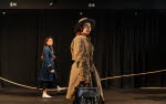 Två personer med flätor och trenchcoats går framåt på en scen.
