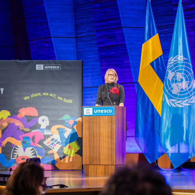 Kvinna som talar för en sittande publik. Unescos textlogotyp på talarstolen.