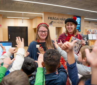 Två kvinnor står vända mot kameran i biblioteksmiljö. Framför dem, med ryggen mot kameran räcker många små barn upp händerna. 