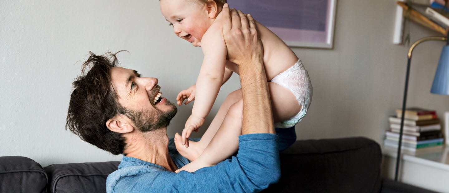 En glad man håller upp en glad bebis i blöja. De ser ut att sitta i deras vardagsrum.