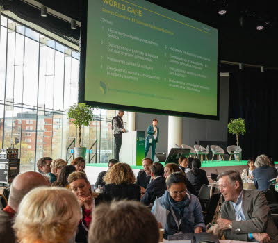 Vy över konferenslokalen och deltagare som sitter i grupper kring bord. På scenen två talare och en stor skärm som visar en presentation. 