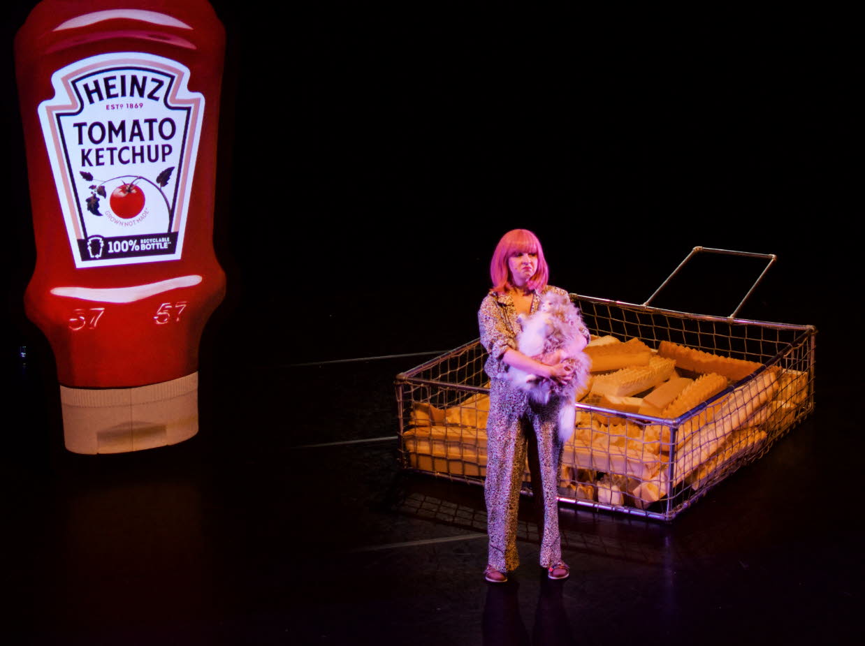 En tjej i rosa hår står på en mörk scen, i bakgrunden syns en förstorad ketchupflaska och förstorade pommes frites..