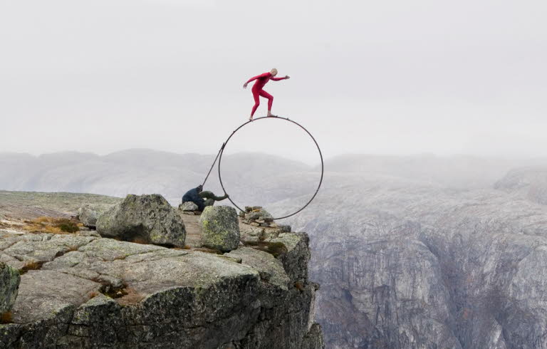 En man klädd i rött står på en stor, smal rund ring precis vid kanten av ett berg.