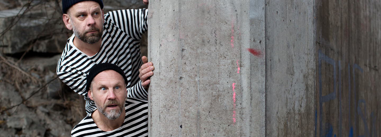 Anders Sparring and Per Gustavsson, dressed in black and white sweaters/prison style, looking out behind a wall.