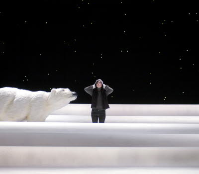 A girl stands on a stage, the background is black and she has snow to her knees, on her right is a polar bear. 