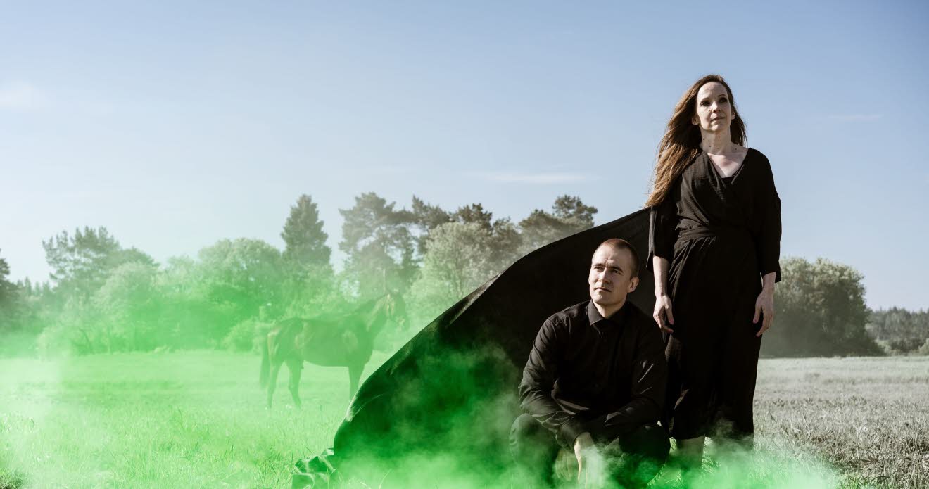 En man och en kvinna i svart klädsel på ett torrt gräsfält med grön rök.