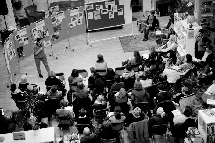  Föreläsning om Women Making HERstory föreläsningssal. Ett 30-tal människor lyssnar till en som står och pratar. Bilden är tagen uppifrån och man ser ingens ansikte tydligt.