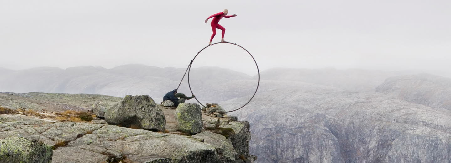 En man klädd i rött står på en stor, smal rund ring precis vid kanten av ett berg.