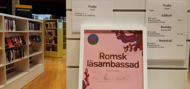 Intyg på att Umeå bibliotek har blivit Romsk läsambassad. Intyget står på ett bord, bokhyllor i bakgrunden. 