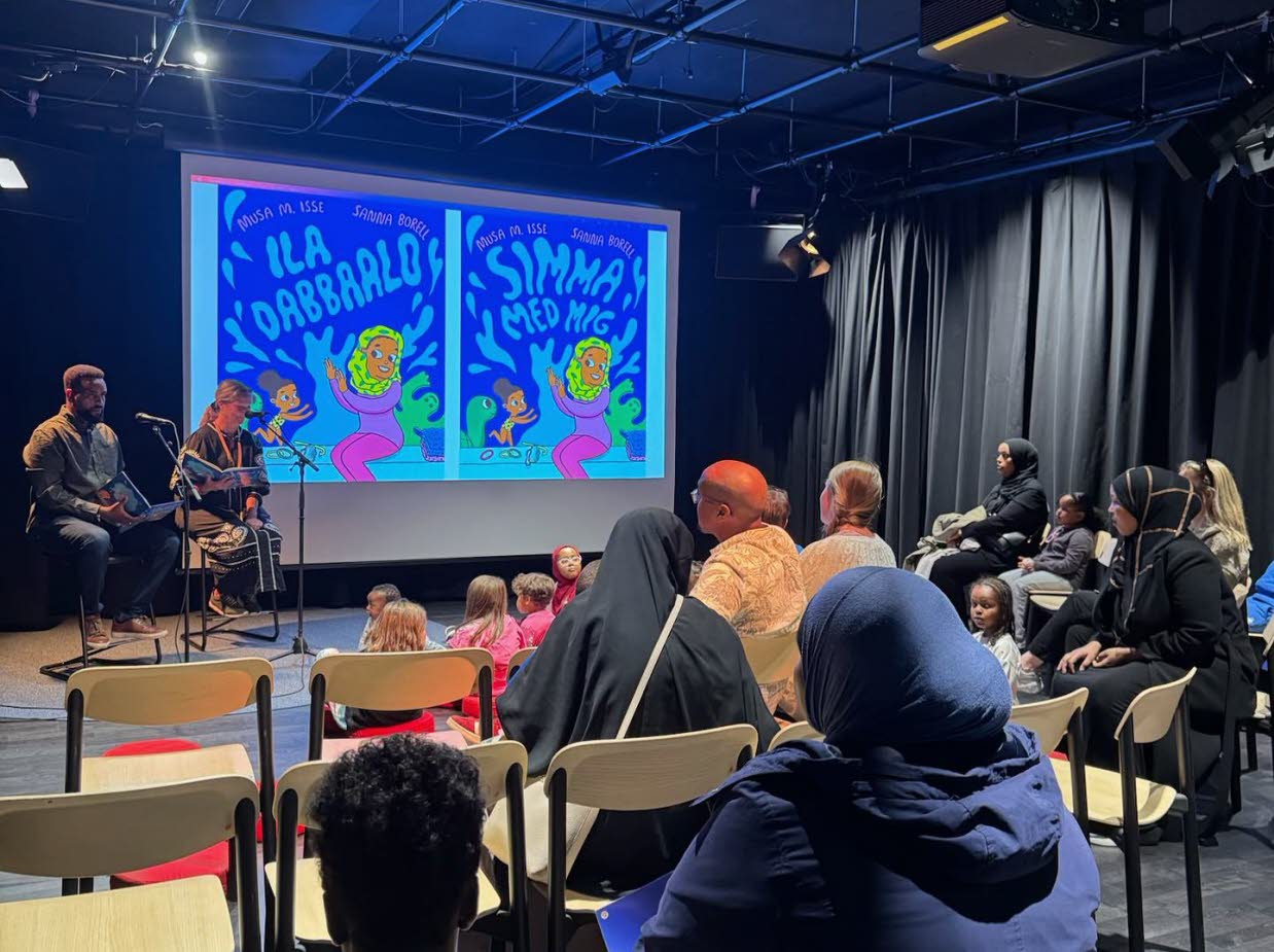 Två personer sitter på en scen och läser framför en publik med barn och vuxna.