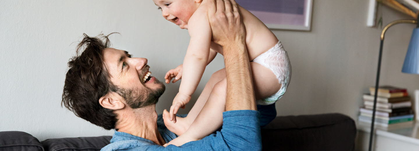 En glad man håller upp en glad bebis i blöja. De ser ut att sitta i deras vardagsrum.