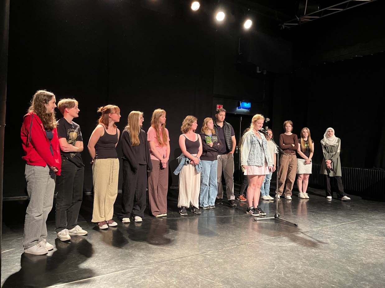 En rad ungdomar står på en scen, en ung kvinna står lite längre fram vid en mikrofon.