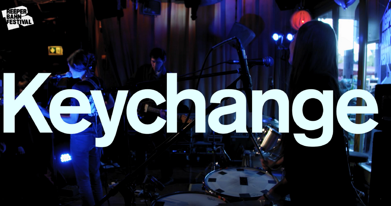 Vit text med Keychange, i bakgrunden ett band som spelar.